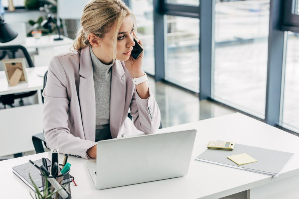 young businesswoman talking by smartphone and using laptop in office